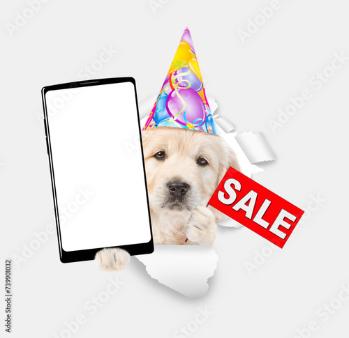 Golden retriever puppy wearing party cap shows big smartphone with white blank screen in it paw and signboard with labeled "sale" through torn white paper hole. Empty free space for mock up, banner