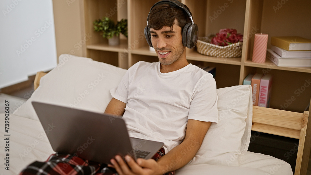 Awake and smiling, a confident young hispanic man enjoys relaxing morning in pajamas, sitting on bed with laptop, listening to music through headphones in his indoor apartment bedroom Awake and smiling, a confident young hispanic man enjoys relaxing morning in pajamas, sitting on bed with laptop, listening to music through headphones in his indoor apartment bedroom