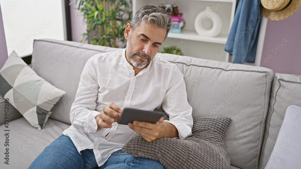 Handsome young hispanic man with grey-haired beard, relaxing at home. concentrated on using touchpad gadget, comfortably sitting on living room sofa, integrating with modern technology.