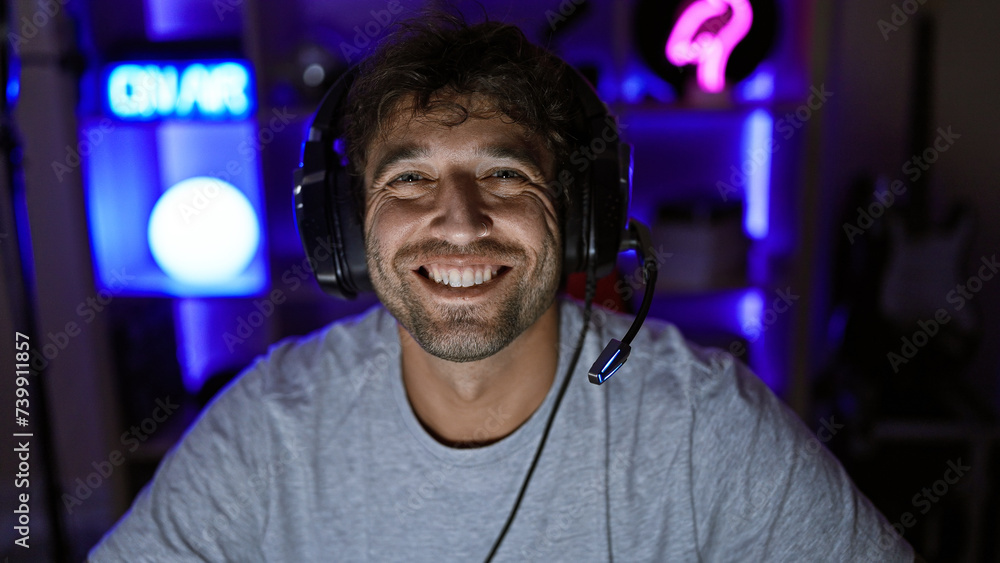 Handsome hispanic man with beard and headphones smiling in a dark gaming room at night Handsome hispanic man with beard and headphones smiling in a dark gaming room at night
