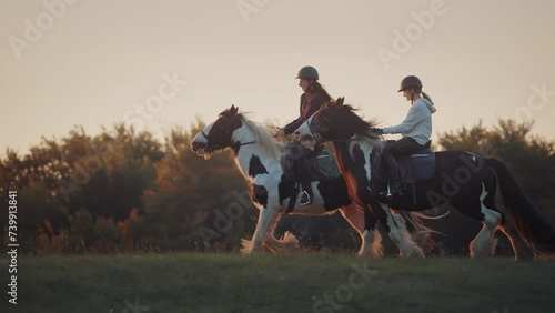 Two young women horseback riding in the green countryside at golden sunset. Riding on a horse in the field. Slowmotion shot. photo