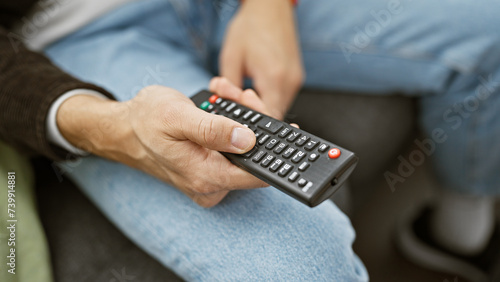 A man relaxing on a couch at home, casually dressed, holding a television remote in a cozy living room setting.