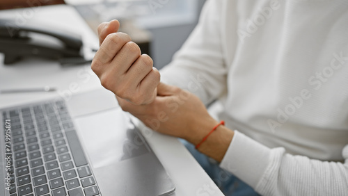 A young adult man with a beard experiences wrist pain while using a laptop in an office setting. photo