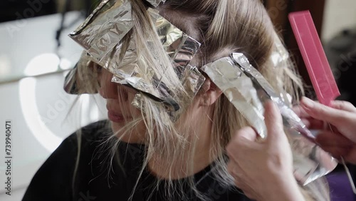  A trendy hair coloring technique is being performed on the young woman at the beauty salon, with the hairdresser applying the color with a brush and wrapping them in foil for processing.