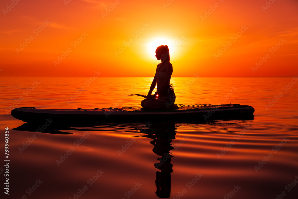 Woman sitting on paddle board at sea with warm bright sunset or sunrise.