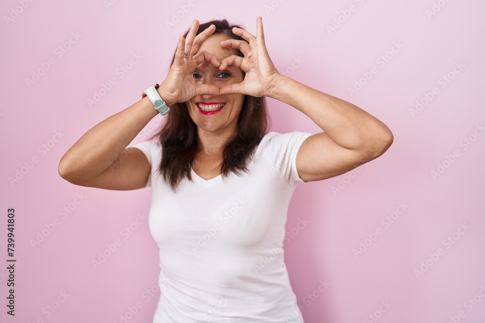Middle age brunette woman standing over pink background doing heart shape with hand and fingers smiling looking through sign Middle age brunette woman standing over pink background doing heart shape with hand and fingers smiling looking through sign