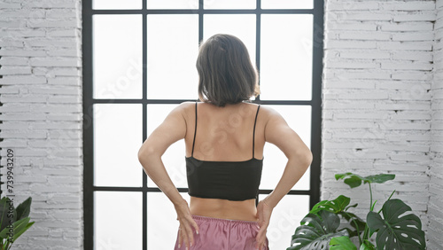 Young  beautiful hispanic woman  throwing us a back view  standing backwards indoors  home lifestyle portrait with short hair  showcased in her apartment s living room interior.