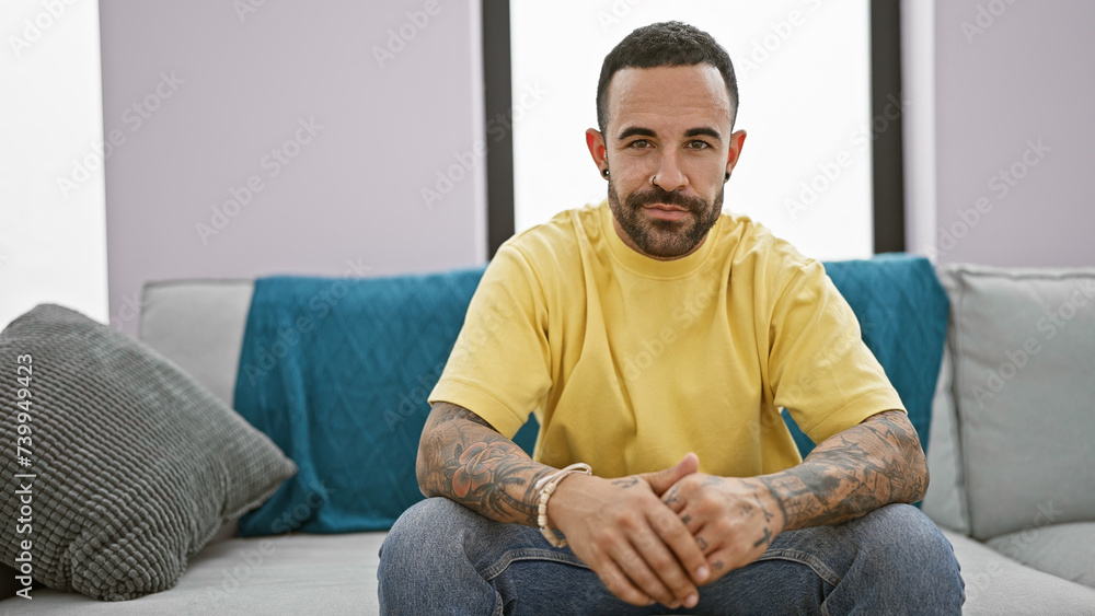 A confident young hispanic man with a beard sits casually in a modern indoor living room setting. A confident young hispanic man with a beard sits casually in a modern indoor living room setting.
