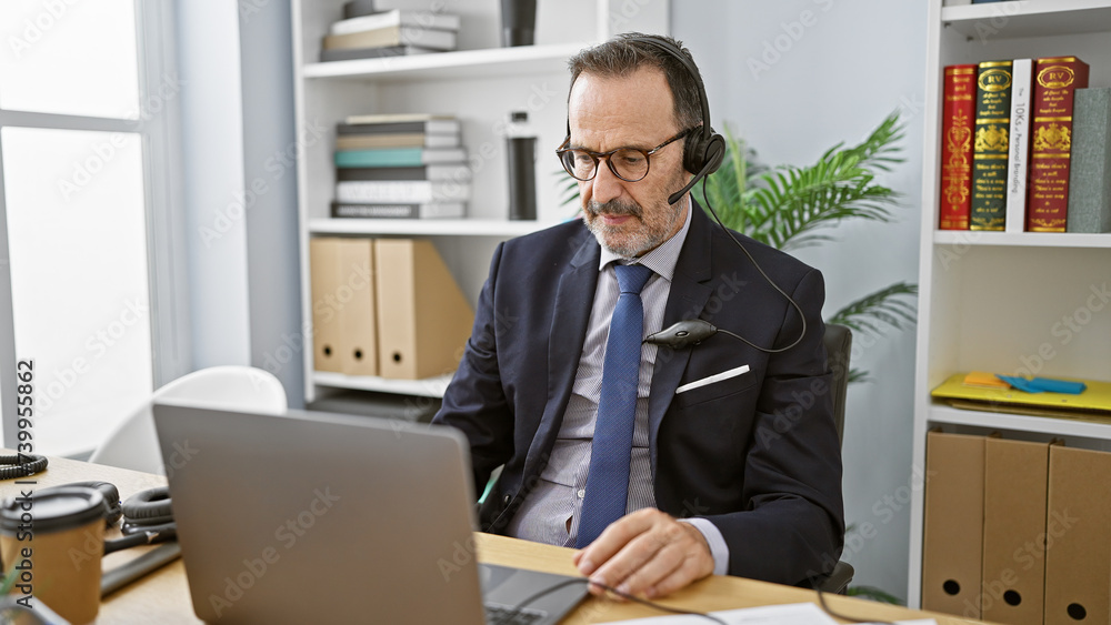 Hispanic middle-aged man with grey hair, focused on business work in the office, professionally speaking to a customer, providing aid through laptop. concentrated, successful worker using headset Hispanic middle-aged man with grey hair, focused on business work in the office, professionally speaking to a customer, providing aid through laptop. concentrated, successful worker using headset