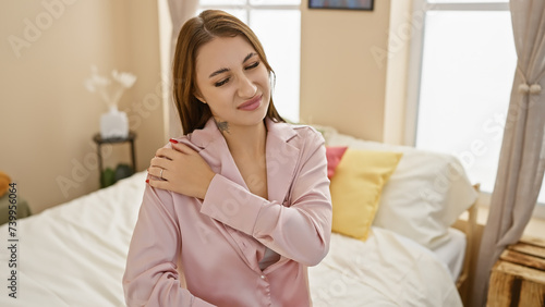 A young beautiful brunette woman in a pink shirt feeling shoulder pain in a well-lit bedroom setting