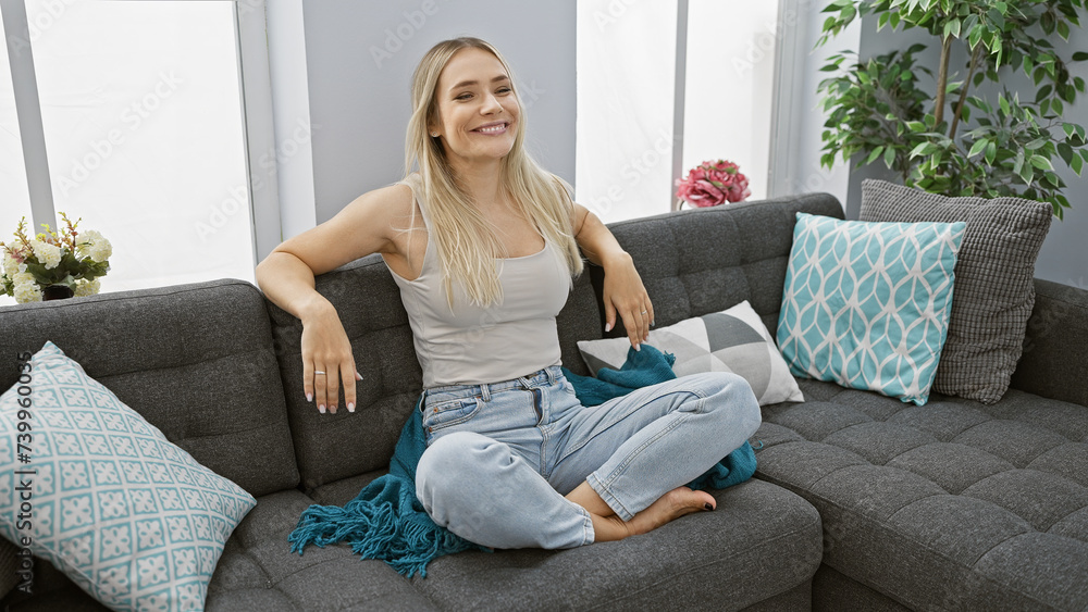 Confident young blonde enjoying relaxation, smiling while sitting comfortably on cozy sofa at home Confident young blonde enjoying relaxation, smiling while sitting comfortably on cozy sofa at home