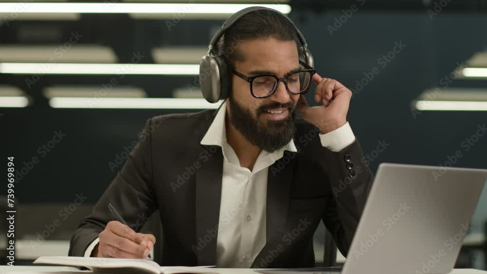 Happy smiling 30s Arabian businessman in headphones looking at laptop watching educational seminar class lesson video call in office Indian business man online negotiations writing notes in copybook Happy smiling 30s Arabian businessman in headphones looking at laptop watching educational seminar class lesson video call in office Indian business man online negotiations writing notes in copybook