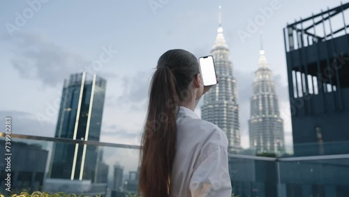 A business woman stares into a phone screen in her hands shooting a video on her phone on the roof of a skyscraper overlooking the big city and the glowing buildings, the lifestyle of the night city.
