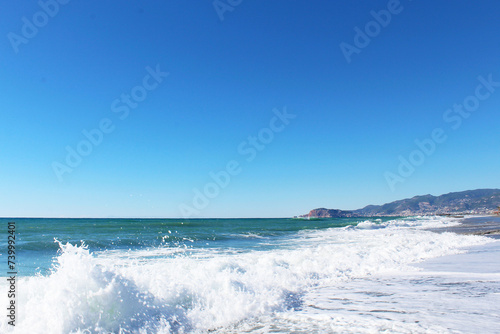 Fototapeta Naklejka Na Ścianę i Meble -  clear blue sky and waves with white foam at Mahmutlar beach in Turkey. mountains in the background