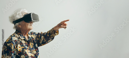 An elderly woman in VR glasses on a light plain background points her finger at an empty space for text.