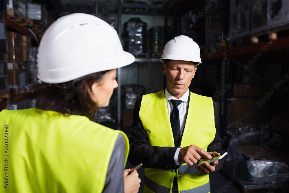 middle aged supervisor in hard hat using smartphone near female employee on blurred background middle aged supervisor in hard hat using smartphone near female employee on blurred background
