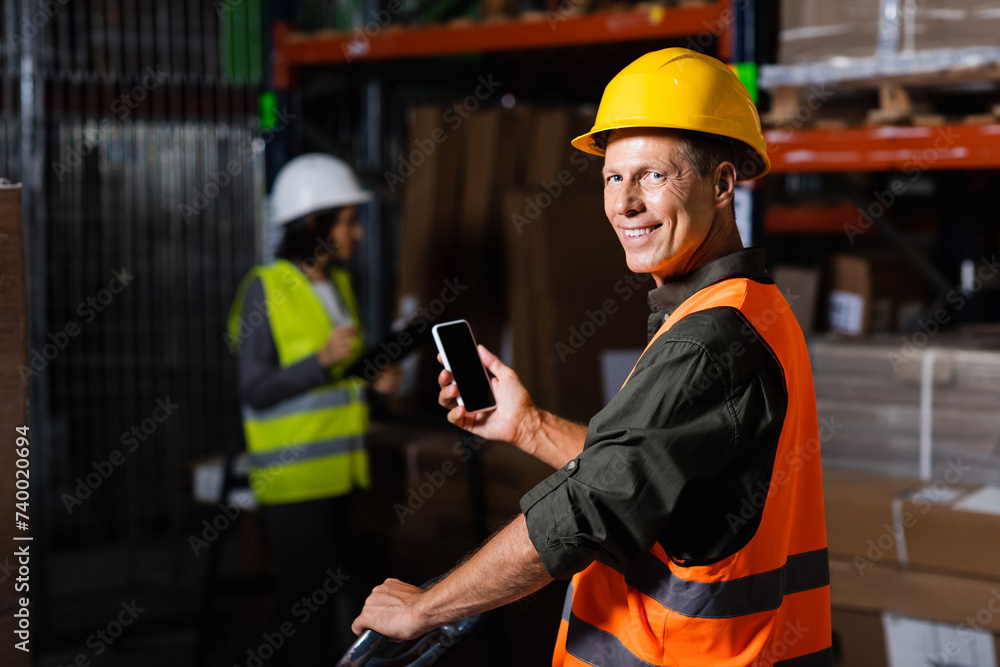 happy supervisor in safety vest using smartphone with employee in background of warehouse happy supervisor in safety vest using smartphone with employee in background of warehouse