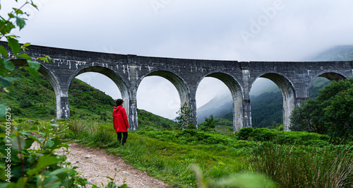 Highland Serenity: A Woman's Journey Through Scotland's Mystical Landscape, Standing Before the Stone Bridge on a Cloudy Day in her Red Coat