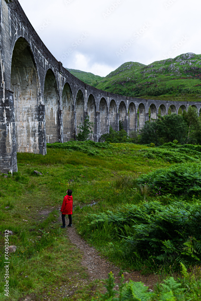 A Woman's Journey Through Scotland's Mystical Landscape, Standing in front of a Stone Bridge on a Cloudy Day in her Red Coat