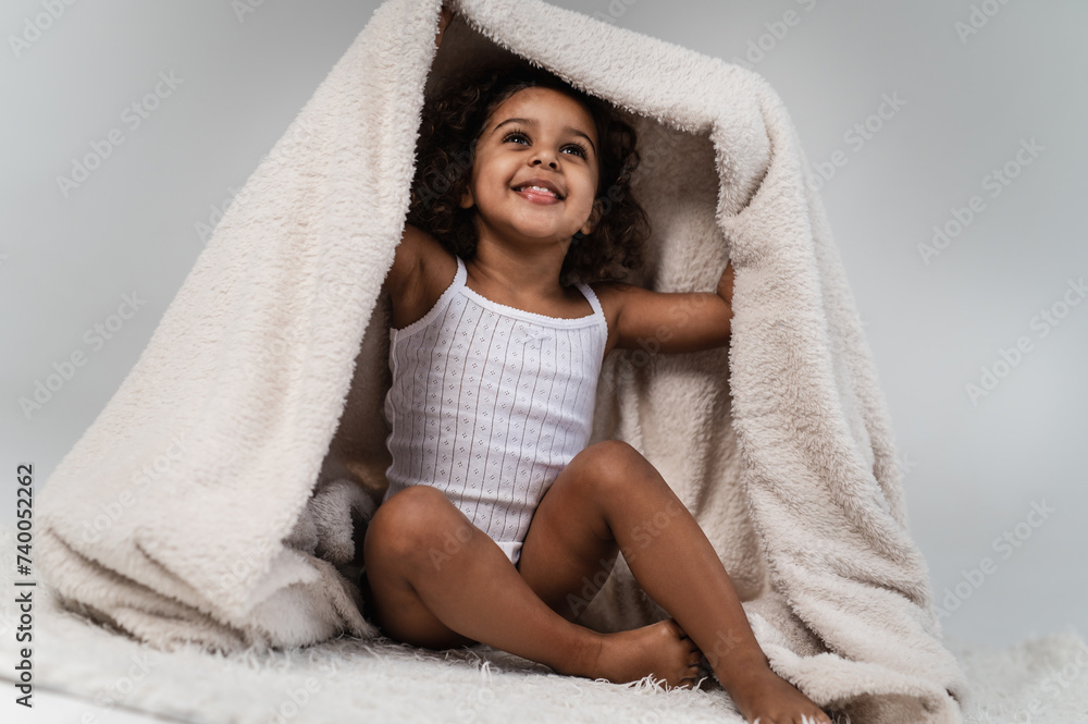 Obraz premium A little cute dark-skinned girl 2 years old plays with a fluffy white blanket on a light plain background. Cozy photos of a cheerful child wrapped in a blanket.