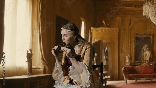 Woman in ancient outfit on background of historic interior. Female in renaissance style dress sitting holding chocolate bar, eating sweets.
