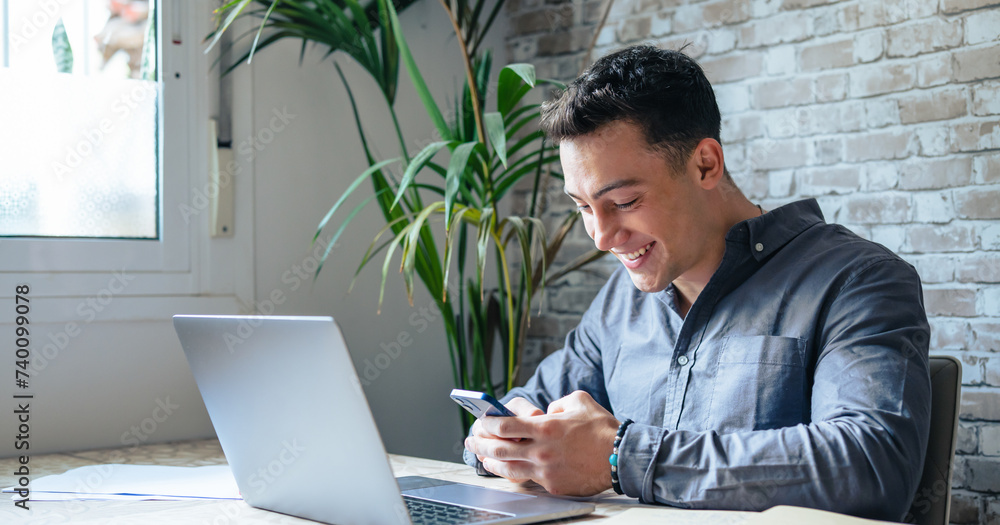 Confident man sitting at desk taking break in work with electronic documents on laptop to make answer telephone call. Smiling young guy freelancer synchronize data between home computer and smartphone Confident man sitting at desk taking break in work with electronic documents on laptop to make answer telephone call. Smiling young guy freelancer synchronize data between home computer and smartphone
