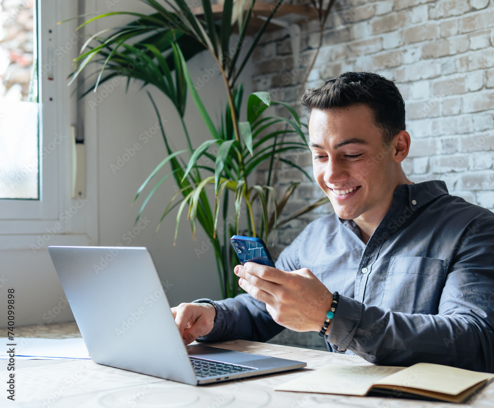 © Daniel - Confident man sitting at desk taking break in work with electronic documents on laptop to make answer telephone call. Smiling young guy freelancer synchronize data between home computer and smartphone