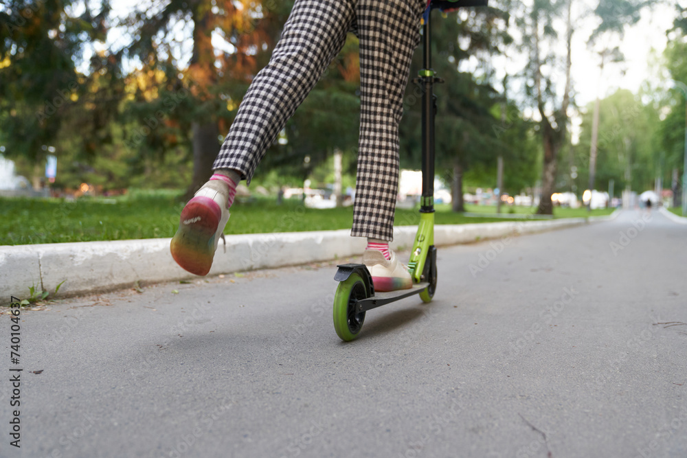 Obraz premium Cropped view from behind of unrecognizable girl in pants riding kick scooters on pavement on summer day, with copy space. Concept of sport, healthy and active lifestyle, eco transport, fun.