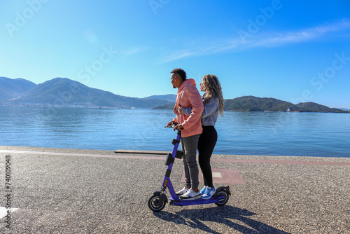 Young latin couple riding a rental e-scooter together along the promenade with the beautiful view on islands. Copy space,