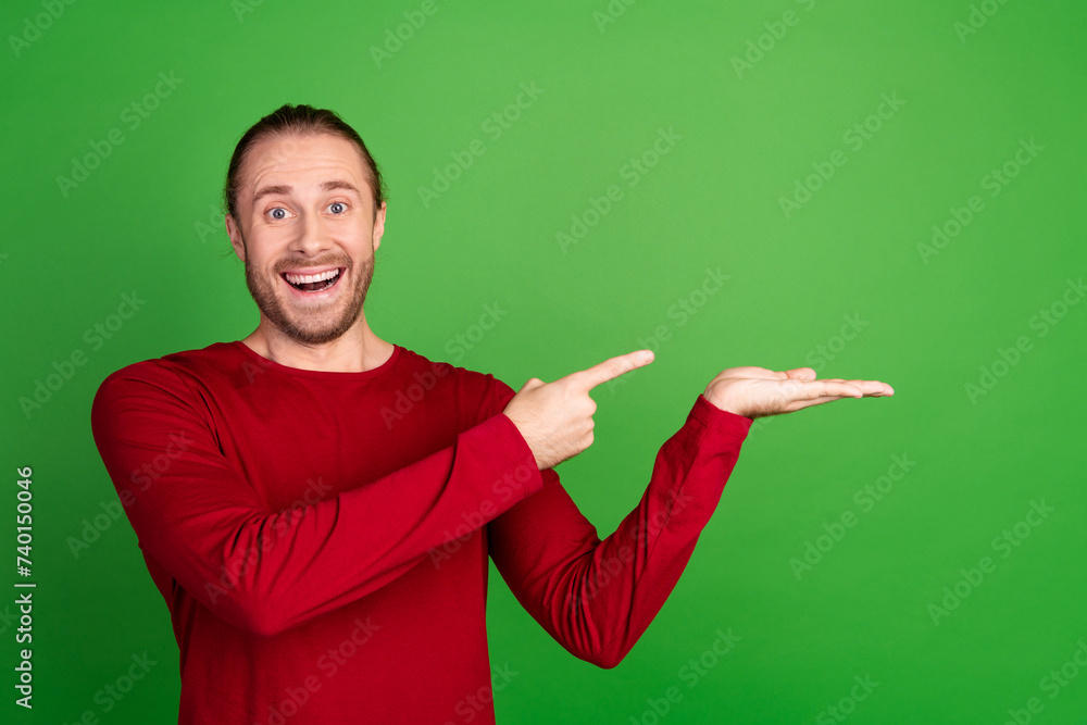 Photo of overjoyed man with stubble dressed red pullover indicating at awesome sale on arm empty space isolated on green color background Photo of overjoyed man with stubble dressed red pullover indicating at awesome sale on arm empty space isolated on green color background
