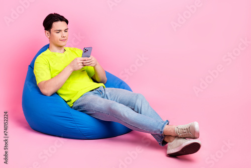 Full length photo of cheerful excited guy wear t-shirt sitting bean bag communicating gadget empty space isolated pink color background