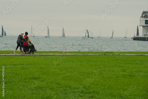 View over green grass of Two women with hats pushing person in a wheelchair on the sidewalk at Vinoy Park looking east towards Tampa Bay, A pier and multiple sailboats in the background. Cloudy day. photo