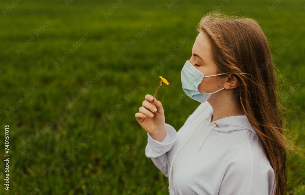 A girl in a respirator sniffs a yellow dandelion. Young blond European woman in a protective mask outdoors. A girl in a respirator sniffs a yellow dandelion. Young blond European woman in a protective mask outdoors.