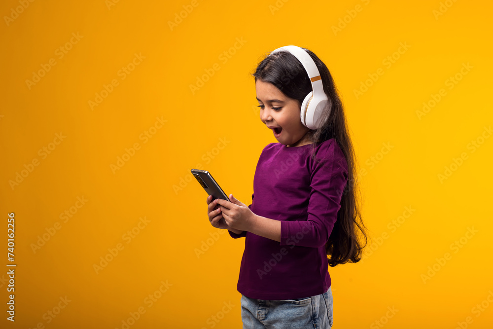 Smiling girl with headphones holding smartprone in hand on yellow background. Lifestyle, leasure and gadget addiction concept Smiling girl with headphones holding smartprone in hand on yellow background. Lifestyle, leasure and gadget addiction concept