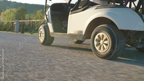 Tracking shot of driving white golf cart on pavement road along the golf course in the countryside, bottom view of cart wheels driving along the road, golf cart delivering players to golf course