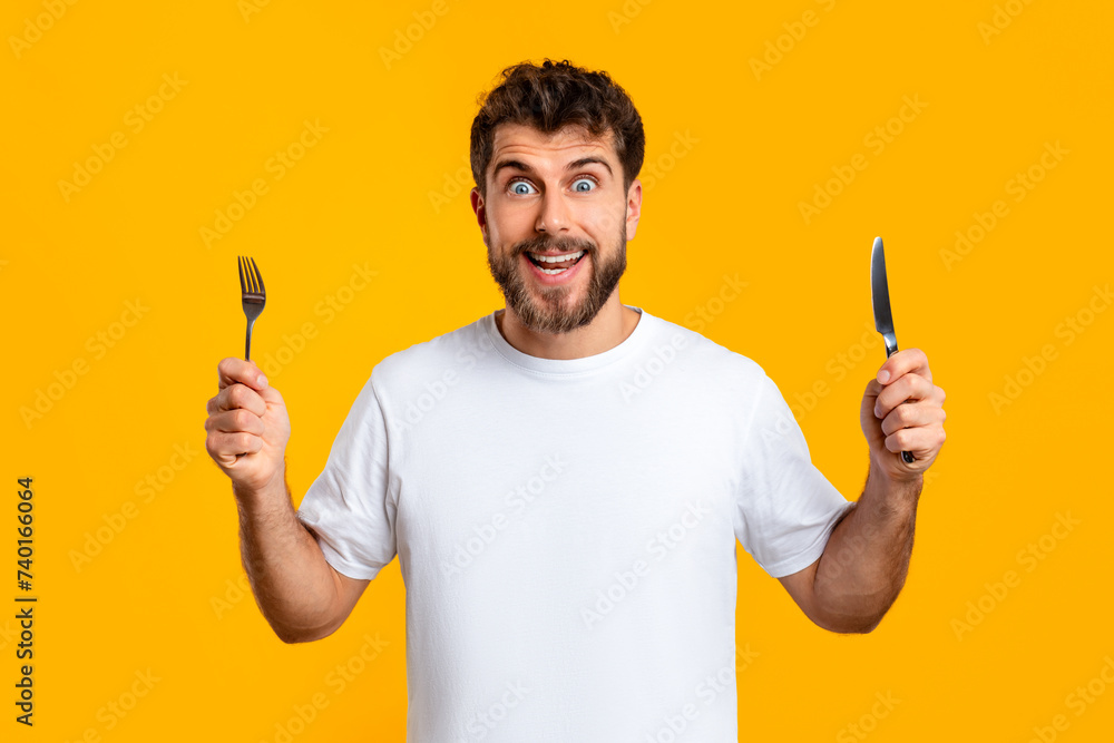 Hungry millennial man holds a knife and fork, yellow backdrop