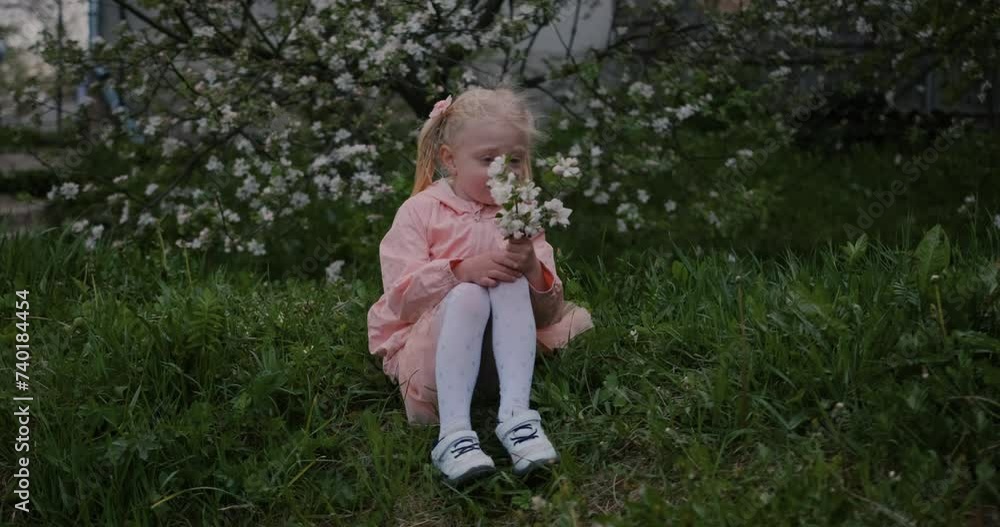 A cute little girl sits with a branch of a blooming apple tree on the grass in the spring garden. A cute little girl sits with a branch of a blooming apple tree on the grass in the spring garden.