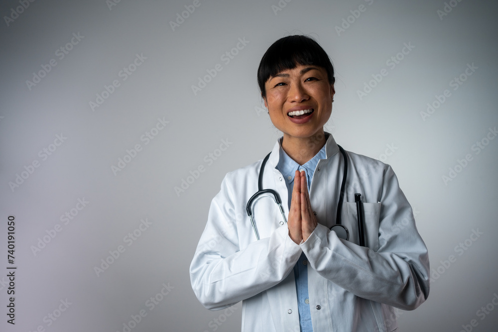 Cheerful female doctor with traditional Japanese greeting isolated on grey background. Positive human face expressions, emotions, attitude. Portrait of a friendly, confident female healthcare doc Cheerful female doctor with traditional Japanese greeting isolated on grey background. Positive human face expressions, emotions, attitude. Portrait of a friendly, confident female healthcare doc