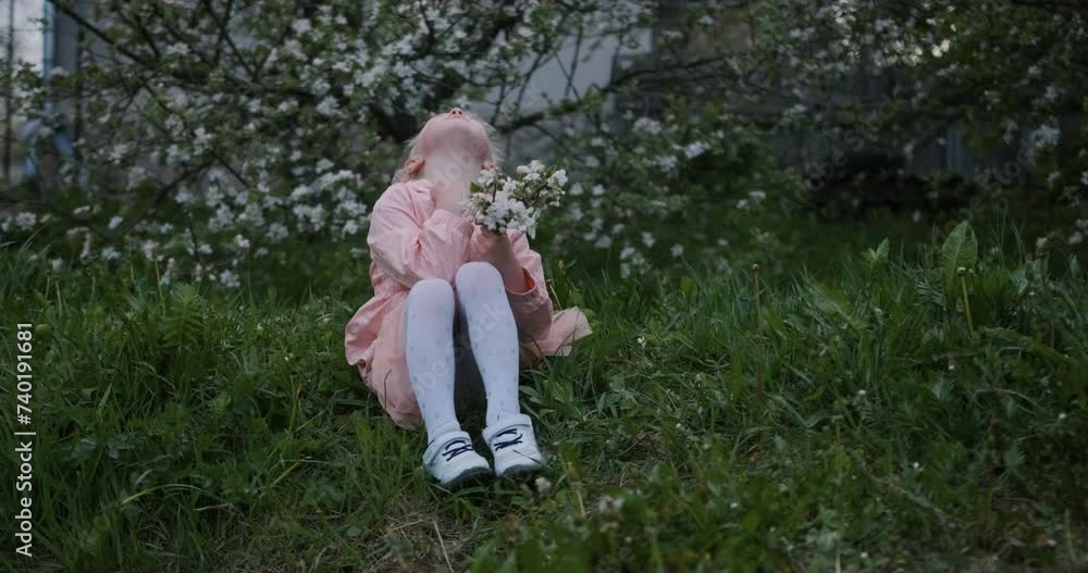Little girl in a pink dress sitting on the grass in a blooming garden and looking up Little girl in a pink dress sitting on the grass in a blooming garden and looking up