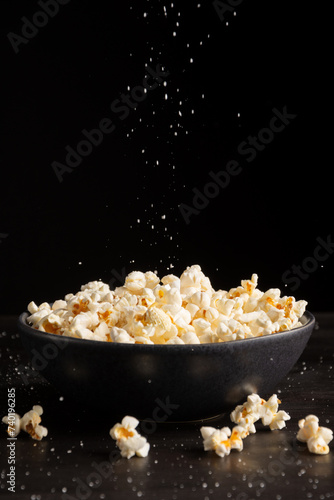 Top view of salt falling into bowl with popcorn on dark table with more popcorn and salt, black background, vertical, with copy space