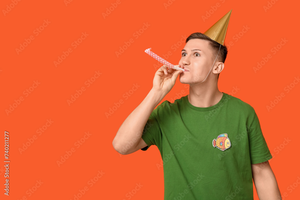Handsome young man with party whistle and paper fish on orange background. April fool's day celebration