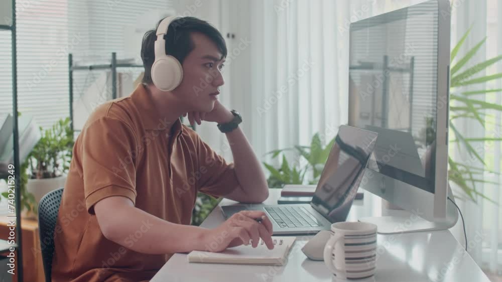 Side view of young Asian tester in headphones working on computer at modern office Side view of young Asian tester in headphones working on computer at modern office