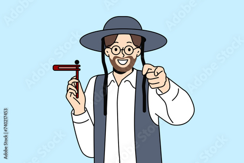 Orthodox jewish man with wooden grager ratchet in hand points finger at screen congratulating you on holiday of hanukkah. Israeli resident preaches judaism invites to celebrate hanukkah with smile