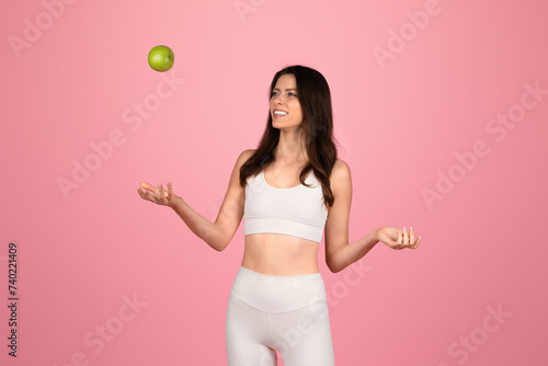 Wall Mural Playful young woman in white fitness attire joyfully tossing a green apple into