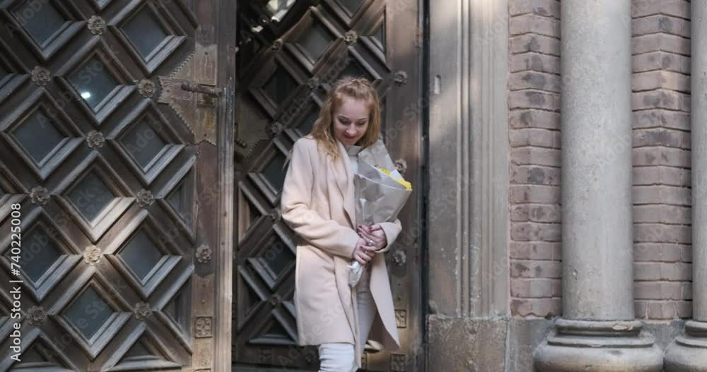 Joyful young blonde woman in beige coat walks out of building with massive wooden doors. Girl holds bouquet of flowers and smiles. Joyful young blonde woman in beige coat walks out of building with massive wooden doors. Girl holds bouquet of flowers and smiles.