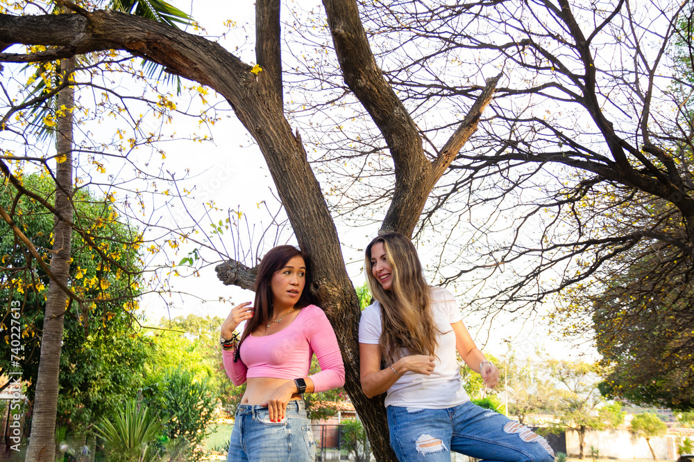 Fototapeta premium two beautiful female friends dressed casual leaning back against a tree looking at each other in a park