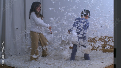 Children having fun in pillow fight with feathers flying in the air, sister battling small brother in speed ramp slow motion at 1000 fps standing in bed