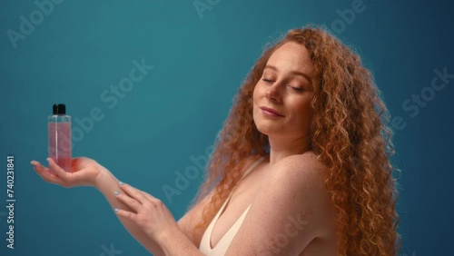 Redhead freckled young plump plus size woman in white lingerie holding palm with bottle of toner. Body positive, skin care and cosmetology.
