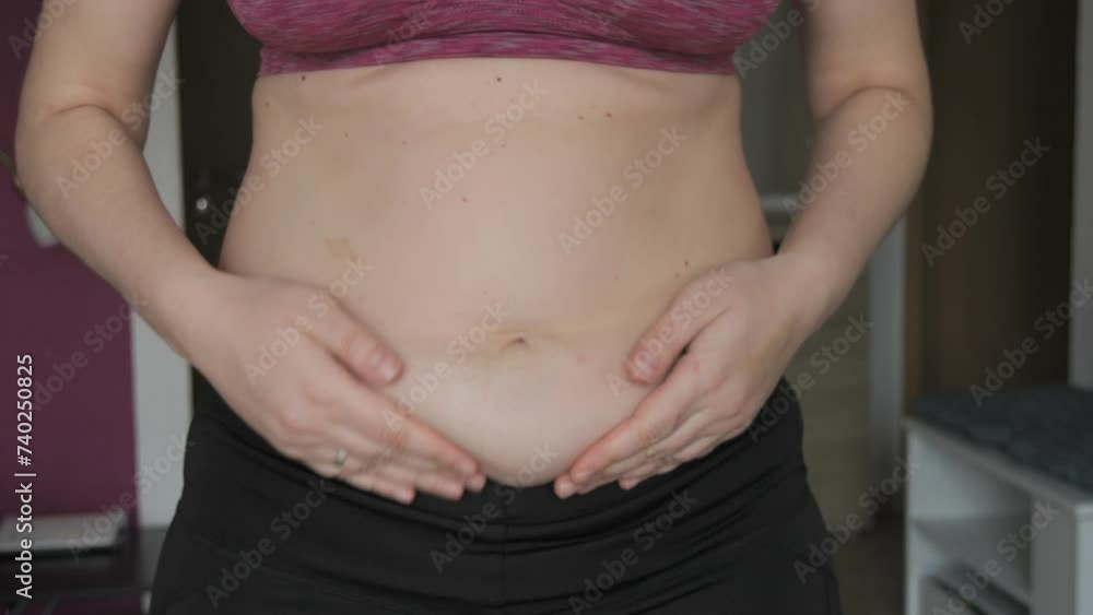 Female belly body positive. Woman touches belly folds in flat slow motion. A small layer of fat on abdomen of woman wearing a sports bra and leggings. Concept of sport, healthy lifestyle or overweight Female belly body positive. Woman touches belly folds in flat slow motion. A small layer of fat on abdomen of woman wearing a sports bra and leggings. Concept of sport, healthy lifestyle or overweight