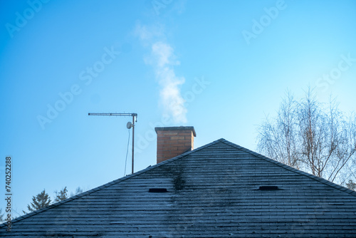 Smoke rises from the chimney on the house. Roof with smoking chimney and trees